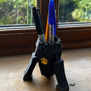 May include: A black, tripod-style desk organiser shaped like a rocket or mechanical structure, featuring a yellow emblem on the front. It holds a black marker, a yellow pencil, and a blue ballpoint pen in its central cylindrical compartment.