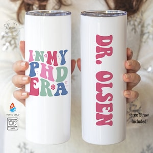 May include: Two white stainless steel tumblers with lids. The tumbler on the left has the text "In My PhD Era" in colorful retro font with stars and flowers. The tumbler on the right has the text "Dr. Olsen" in pink retro font.