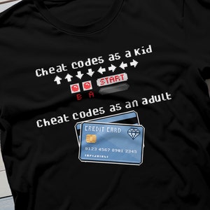 Pode incluir: Camiseta preta com um design pixelado que diz "Cheat codes as a kid" com setas para cima e para baixo e um botão "START". Abaixo, diz "Cheat codes as an adult" com um cartão de crédito pixelado com o número "0123 4567 8981 2345".