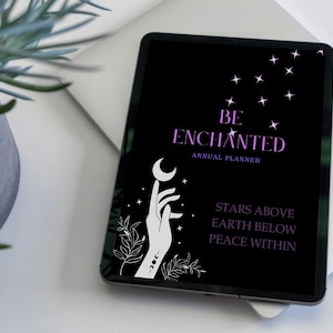 May include: A black tablet with a white hand holding a crescent moon. The text "BE ENCHANTED ANNUAL PLANNER" is in purple on the screen. The text "STARS ABOVE EARTH BELOW PEACE WITHIN" is also in purple.