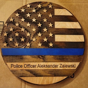 May include: A round wooden sign with a distressed American flag design. The flag has a blue stripe with a thin white line above it. The sign has the text "Police Officer Aleksander Zalewski" below the flag.