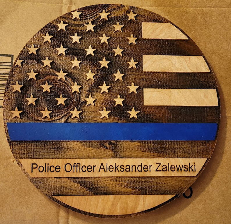 May include: A round wooden sign with a distressed American flag design. The flag has a blue stripe with a thin white line above it. The sign has the text "Police Officer Aleksander Zalewski" below the flag.