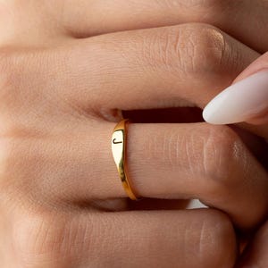 Può includere: Un anello dorato con una piccola lettera "J" incisa è mostrato su un dito. L'anello ha una superficie liscia e lucida e un design semplice. Lo sfondo è sfocato, concentrando l'attenzione sul gioiello. L'anello è probabilmente d'oro.