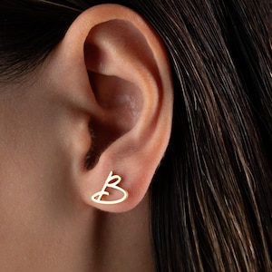 Peut inclure: Une paire de boucles d'oreilles dorées en forme de lettre "B" cursive et délicate. Les boucles d'oreilles sont petites et minimalistes, conçues pour être portées dans les lobes d'oreille. Les boucles d'oreilles sont portées.