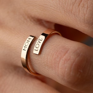 May include: A gold-colored ring with the words "MOM" and "LOVE" engraved on it. The ring is wrapped around a finger, showcasing its design. The ring's simple yet elegant design makes it a perfect gift.