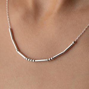 May include: A delicate silver necklace featuring a series of small, cylindrical beads and accents. The necklace has a curved design and is suspended from a fine chain. The jewelry is shown against a neutral background.