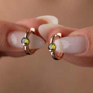 May include: A pair of rose gold hoop earrings, each featuring a small, round, olive-green gemstone. The earrings are held up by a person's hands, showcasing the delicate design and color contrast. The hoops are small and hug the earlobe.