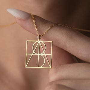 May include: A gold pendant necklace with a geometric design of lines, circles, and a square. The pendant hangs from a delicate gold chain. The necklace is being held up by a person's finger.