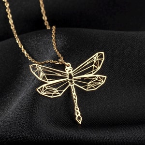 May include: A gold necklace with a geometric dragonfly pendant. The necklace is on a black fabric background.