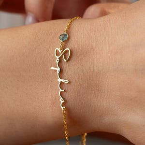 May include: A delicate gold bracelet with the name "Sophia" in cursive script. The bracelet features a small, light blue gemstone and is worn on a wrist. The jewelry is a personalized accessory.