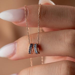 May include: A delicate gold necklace featuring two interlocking rings. One ring is adorned with dark blue gemstones, the other with light blue gemstones. The necklace is held by a person with manicured nails, showcasing the jewelry's design.