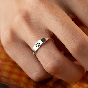 Puede incluir: Un anillo de plata con un diseño de huella de pata. El anillo es curvo y presenta una pequeña huella de pata negra en la parte delantera. El anillo se muestra en un dedo.
