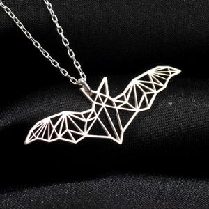 May include: A silver-colored necklace with a geometric bat pendant. The bat design is made of interconnected lines, creating a polygonal shape. The necklace hangs on a delicate chain against a black background.