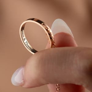 Könnte beinhalten: Ein roségoldener Ring mit der Gravur "Anna & Thomas". Der Ring hat ein filigranes Muster am Band. Der Ring wird zwischen zwei Fingern vor einem unscharfen Hintergrund gehalten.