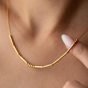 May include: A delicate gold necklace featuring a series of small, cylindrical and spherical beads. The necklace is worn around the neck, with the chain and beads appearing to be made of a shiny, yellow metal. The design is simple and elegant.