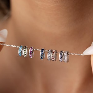 Könnte beinhalten: Eine silberne Halskette mit mehreren kleinen, farbigen Ringen. Jeder Ring ist mit kleinen, funkelnden Edelsteinen in verschiedenen Farben, darunter Blau, Grün, Rosa und Orange, verziert. Die Halskette wird von einer Hand gehalten.