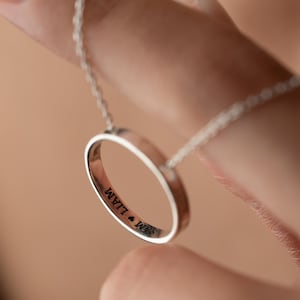 Könnte beinhalten: Eine silberfarbene Halskette mit einem Anhänger in Form eines kreisförmigen Rings. Der Ring trägt die eingravierten Namen "SAM" und "LIAM". Die Halskette wird von einer feinen Kette gehalten. Der Hintergrund ist unscharf.