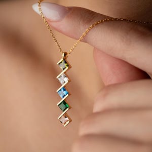 May include: A gold-colored necklace with a series of colorful, square-cut gemstones. The gemstones are arranged in a zig-zag pattern, with colors including green, white, blue, and pink. The necklace is held up by a hand.