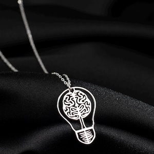 Op de afbeelding: Een zilveren ketting met een hanger in de vorm van een gloeilamp. De gloeilamp heeft een uitgesneden ontwerp van een brein erin.