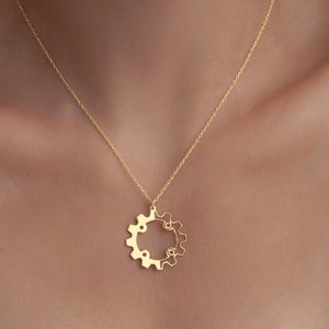 Op de afbeelding: Een gouden ketting met een tandwielvormige hanger. De hanger is gemaakt van twee in elkaar grijpende tandwielen.