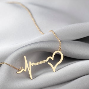 Peut inclure: Collier doré avec un motif de battement de cœur et un pendentif en forme de cœur. Le collier comprend la date "27.08.2021" et repose sur une surface en satin gris. Un bijou délicat.