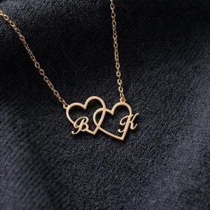 Op de afbeelding: Een goudkleurige ketting met twee verbonden hartvormige hangers. Elk hart heeft een letter, met de letters "B" en "K" zichtbaar. De ketting is delicaat en de achtergrond is een donkere, getextureerde stof.