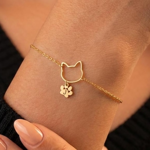 May include: A gold-colored bracelet featuring a cat face outline and a paw print charm. The delicate chain bracelet is worn on a wrist, showcasing the jewelry's design and detail. The bracelet is a charming accessory for cat lovers.