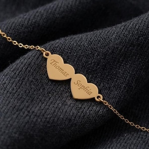 May include: A gold-colored necklace with two heart-shaped pendants. Each heart is engraved with a name: "Thomas" and "Sophia". The necklace chain is delicate, and the background is a dark, textured fabric.