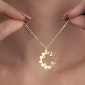 Puede incluir: Un collar de cadena dorada con un colgante en forma de engranaje. El colgante es un solo engranaje con una abertura circular en el centro.