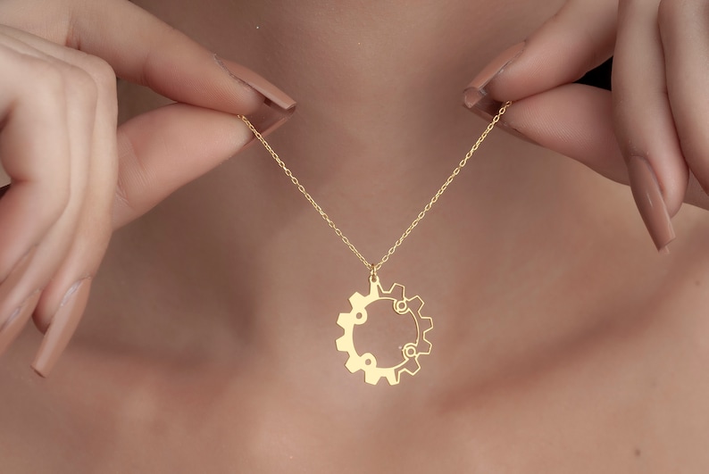 Puede incluir: Un collar de cadena dorada con un colgante en forma de engranaje. El colgante es un solo engranaje con una abertura circular en el centro.