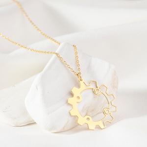 May include: A gold-colored necklace featuring a gear pendant. The pendant has a detailed, openwork design with a circular shape and intricate cutouts. The necklace is displayed on a white stone against a white fabric background.