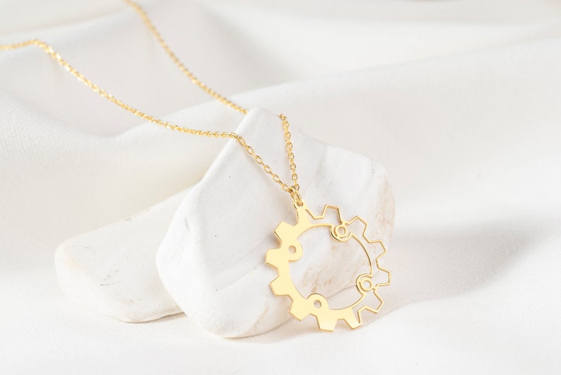 May include: A gold-colored necklace featuring a gear pendant. The pendant has a detailed, openwork design with a circular shape and intricate cutouts. The necklace is displayed on a white stone against a white fabric background.