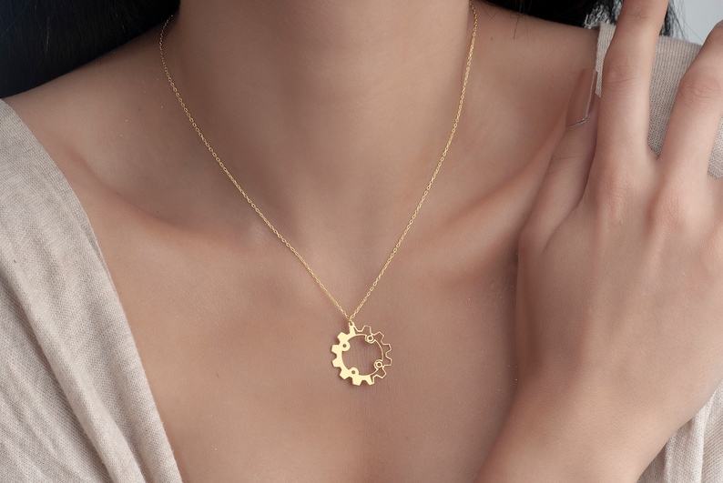 Puede incluir: Un collar de cadena de oro con un delicado colgante de engranaje. El engranaje es un c&iacute;rculo con peque&ntilde;os dientes alrededor del borde.