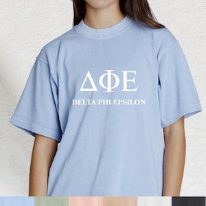 May include: Light blue t-shirt with the Greek letters ΔΦΕ and the words DELTA PHI EPSILON printed in white. The shirt has short sleeves and a crew neck.