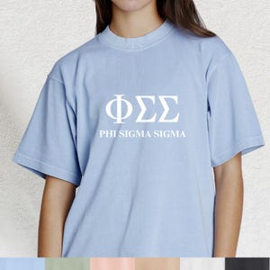 May include: A pale blue t-shirt featuring the Greek letters ΦΣΣ and the words PHI SIGMA SIGMA in white. The shirt has a crew neck and short sleeves.