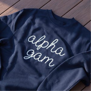 May include: Navy blue sweatshirt with the words "alpha gam" embroidered in white cursive script across the front. The sweatshirt is made of a soft material and has a crew neck.