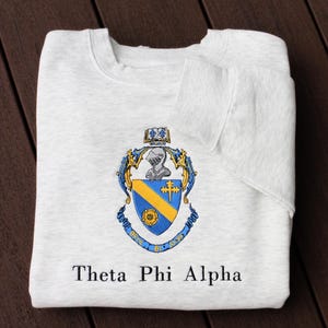 May include: A white sweatshirt with a blue and yellow crest and the text "Theta Phi Alpha" printed on the front.