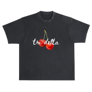 May include: Dark gray t-shirt featuring a watercolor illustration of two red cherries with green stems. The words "tri delta" are written in white script below the cherries. The shirt has a classic crew neck and short sleeves.