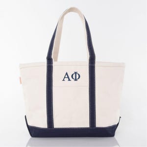 May include: Cream-colored canvas tote bag with navy blue accents. Features navy blue handles and a navy blue bottom. The bag has the embroidered letters AΦ in navy blue on the front. A classic and durable tote bag.