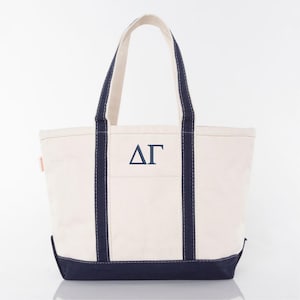 May include: Cream-colored canvas tote bag with navy blue accents. Features navy blue handles and a navy blue bottom. The front of the bag displays the Greek letters ΔΓ in navy blue. A practical and stylish accessory.