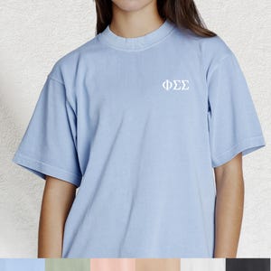 May include: A pale blue short-sleeved t-shirt featuring the Greek letters ΦΣΣ in white on the chest. The top has a crew neck and a loose fit. The material looks soft and slightly faded.