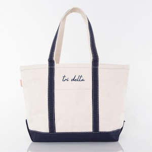 May include: Cream-colored tote bag with navy blue accents. The bag features navy blue handles and a navy blue base. The words "tri delta" are embroidered in navy blue on the front of the bag. The bag is made of a sturdy canvas material.