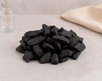 May include: A pile of dark black decorative rocks. The rocks have rough, irregular shapes and varying sizes. The background includes a clear glass vase with dried flowers and a neutral-colored cloth.