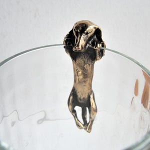May include: A bronze-colored dog figurine, possibly a dachshund, hangs over the rim of a clear glass. The dog's head and body are detailed, with a textured finish. The figurine appears to be a decorative item, possibly a glass charm or ornament.
