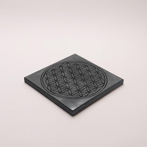 Puede incluir: Una piedra cuadrada, gris oscuro, con un detallado patrón grabado de la Flor de la Vida. El diseño geométrico está centrado en la piedra, sobre un fondo rosa pálido. La piedra parece lisa.