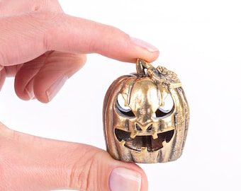 May include: A small, metallic gold-colored pumpkin with carved-out eyes, nose, and mouth. The pumpkin has a stem and a leaf detail. The Halloween-themed decoration is held against a white background.