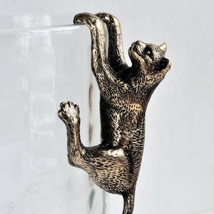 May include: A gold-colored metal cat figurine is clinging to the side of a clear glass mug. The cat is depicted in a playful pose, with its paws gripping the mug and its tail curled around the side.