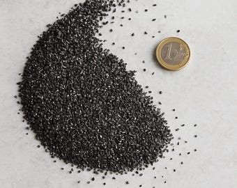 May include: A pile of small, dark gray granules with a 1 Euro coin for size comparison. The granules are scattered across a light gray surface. The coin is gold and silver in color.