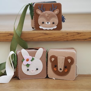 May include: Three felt baby books with animal faces: a deer, a bunny, and a bear. The deer book is brown with a white collar and antlers. The bunny book is tan with a pink flower. The bear book is tan with a brown face.