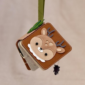 May include: A handmade felt baby book with a deer design on the cover. The book is brown, tan, and white, with a green ribbon for hanging. The deer has a white bib and gray antlers. A small black turtle charm hangs from the book.